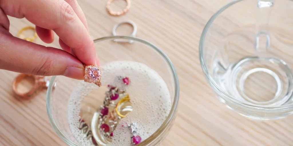 A hand holding a ring above a glass bowl filled with soapy water and jewelry inside, showing how to clean artificial jewelry effectively at home.