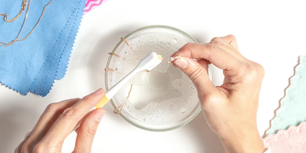 Hands showing how to clean oxidised jewellery with a toothbrush over a bowl of soapy water, flanked by colorful cloths.