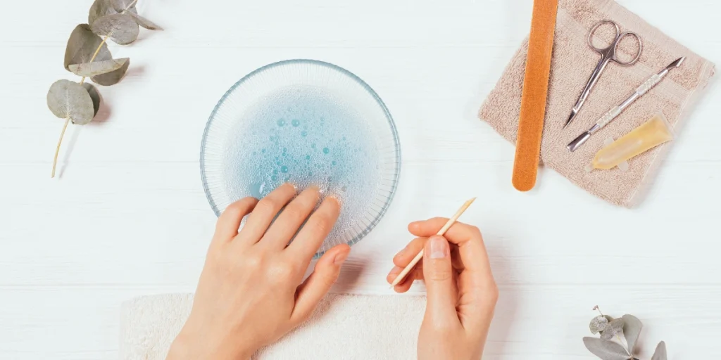 Person's hands cleaning jewelry with tools and a bowl of soapy water on a white surface.