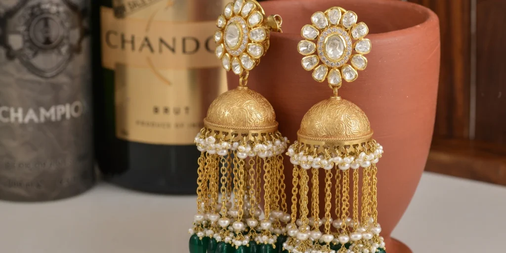 Gold and pearl jhumka earrings with floral designs, set against a backdrop of wine bottles and a clay pot. (What jewellery to wear with lehenga)