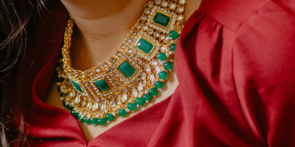 Close-up of a woman wearing an ornate gold necklace adorned with emerald and clear stones, contrasted against a red blouse.