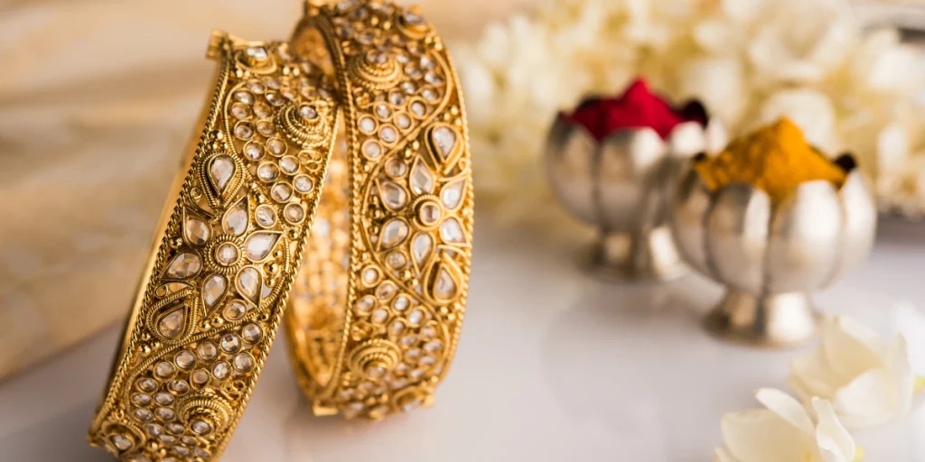 Two ornate golden bangles adorned with intricate patterns and stones, beside silver bowls filled with colorful powders and white flowers. (What jewellery to wear with lehenga)
