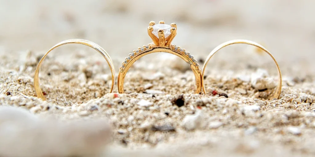 Three gold rings partially buried in sandy beach, with a central engagement ring adorned with a sparkling gemstone.