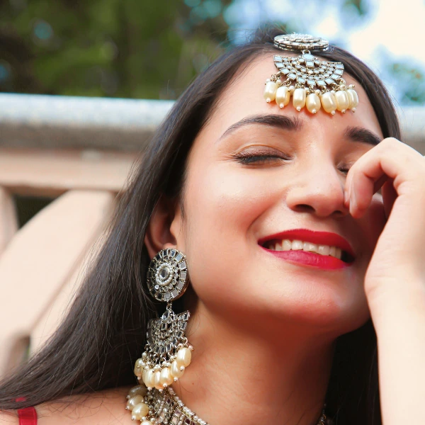A woman wearing intricate silver jewelry and a red outfit poses with her hand near her face, set against a blurred outdoor background.